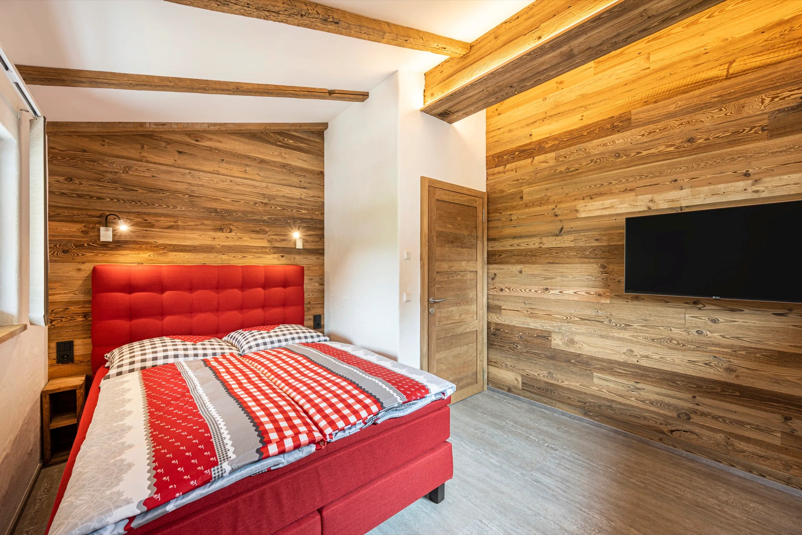 Schlafzimmer mit Altholzwand, rotem Bett und Fernseher